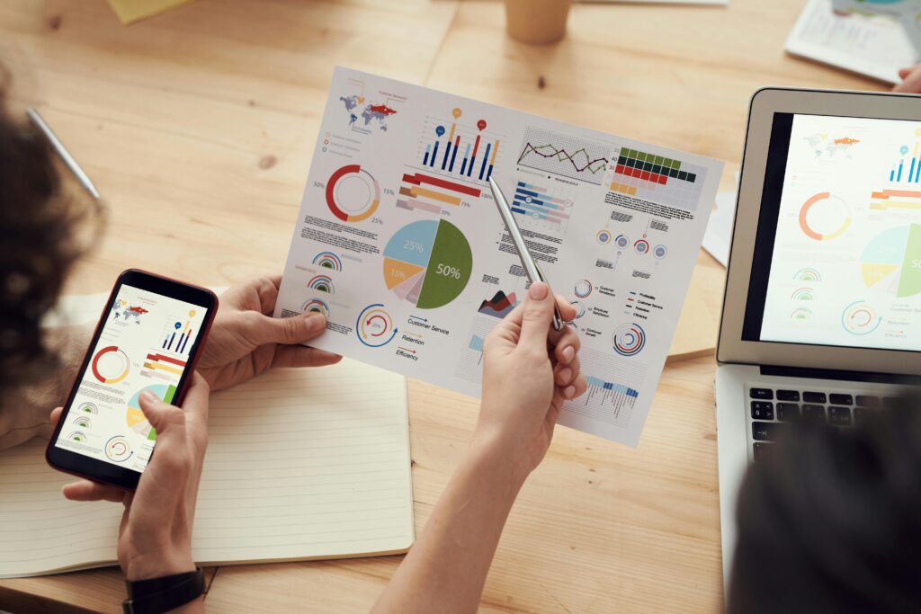 Two people are looking at a report filled with charts and graphs. One person is holding a phone displaying similar data, and another person is using a laptop in the background. A hand points to a pie chart on the report which shows 50%