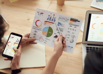 Two people are looking at a report filled with charts and graphs. One person is holding a phone displaying similar data, and another person is using a laptop in the background. A hand points to a pie chart on the report which shows 50%